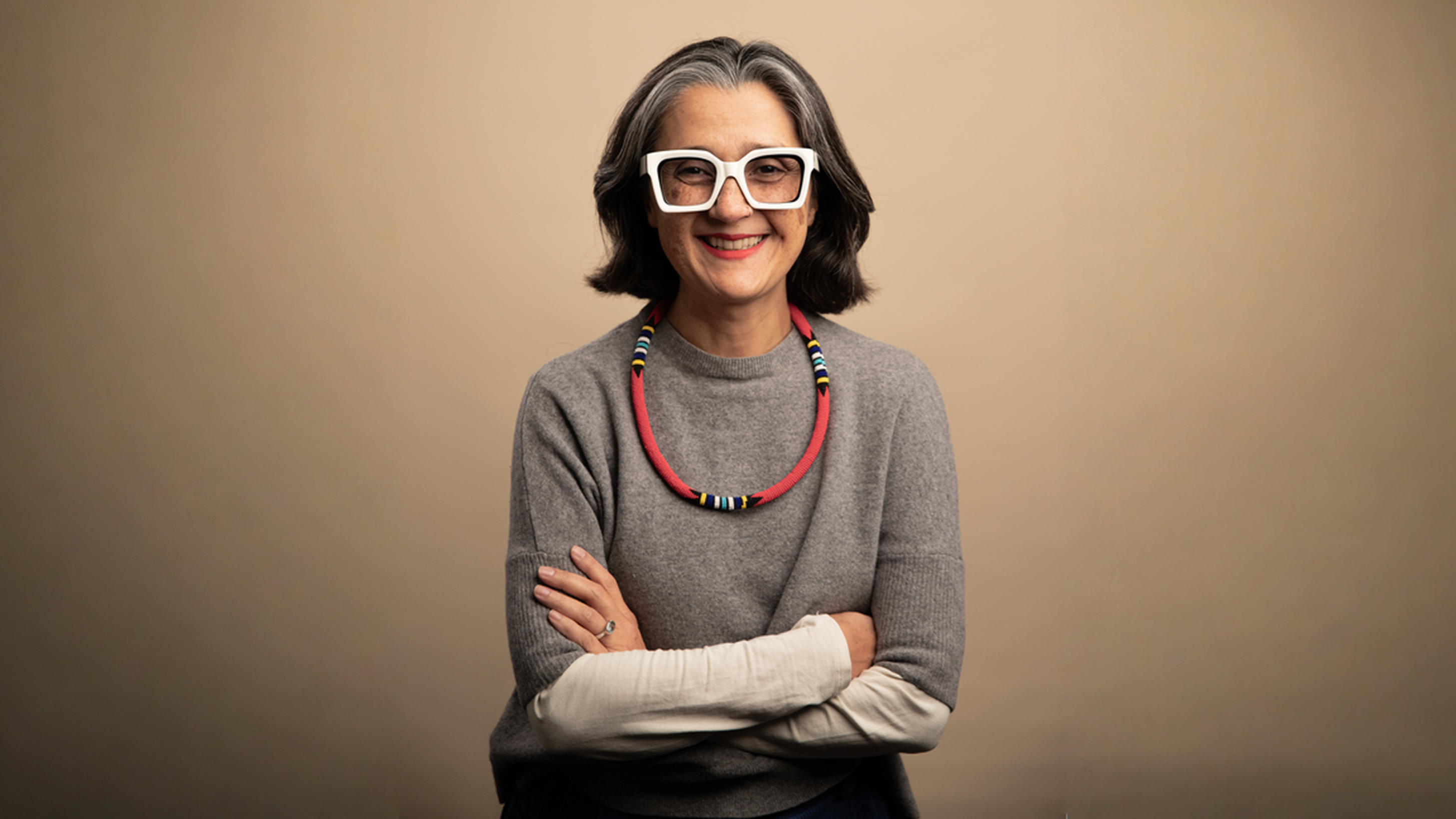 A photo shoot for a woman posing for the camera with neutral colour background. She has medium-lenght grey hair, wearing big white frame glasses and smiling.